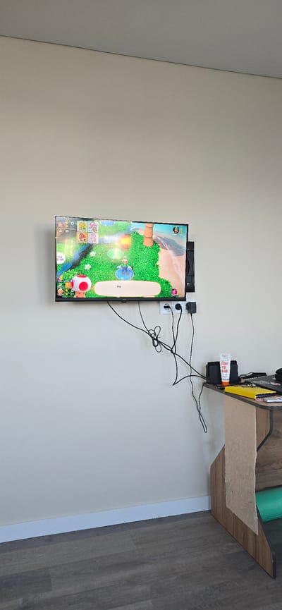 The image shows a wall-mounted television displaying a video game. The game appears to be a party game with multiple characters, including Mario and Toad. The TV is connected to a power outlet with several cables. To the right of the TV, there is a small shelf with a black box, a tube of cream, and some keys. A notebook is also visible on the shelf. The wall is painted a light beige color, and the floor is a dark wood laminate. The image is taken indoors, likely in a living room or entertainment area. There is no specific information in the image to indicate it is located in Bryansk, Russia.