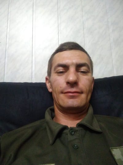 The media file is a close-up, slightly angled selfie of a man. He is lying down, with his head resting on a dark surface, possibly a couch or bed. He is wearing a dark green collared shirt with buttons. A camouflaged patch is visible on the left side of his shirt.

The man's face is turned towards the camera. He has short, dark hair and light skin with some freckles. His expression appears neutral or slightly relaxed. The background consists of a white, paneled wall with horizontal lines.

The lighting suggests it is daytime, possibly indoors. There are no obvious weather cues. The overall impression is informal and personal. No text is visible in the image.