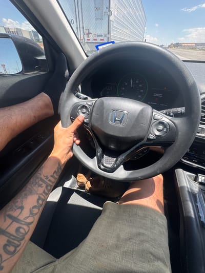 The image shows a first-person perspective of a person driving a car. The driver's left foot, wearing a black sock, is resting on the dashboard. Their right hand is on the steering wheel, which has Honda branding and various control buttons. The dashboard displays speed and other vehicle information, including an "Airbag System (SRS) Problem" warning light. The driver is wearing shorts, and tattoos are visible on their left forearm. Outside the car, a large truck and other vehicles are visible, along with buildings and a bright sky, suggesting an urban or industrial area. The scene appears to be on a sunny day. The specific location in Socorro, United States, is not discernible from the visual information provided.