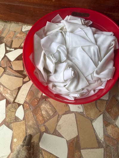 The image, taken in Tela, Honduras, shows an overhead view of a red plastic laundry basket containing numerous folded and rolled white fabric items, possibly sheets or tablecloths, with a slight sheen. The basket features integrated handles on its rim and rests on a floor covered in an irregular mosaic pattern of beige, brown, and off-white ceramic tile shards. In the upper background, a dark brown wooden surface is partially visible. In the lower left foreground, a small, textured, light-colored object, resembling a mop head or animal fur, is visible on the tiled floor. There are no people present or explicit actions depicted beyond the static arrangement of objects.