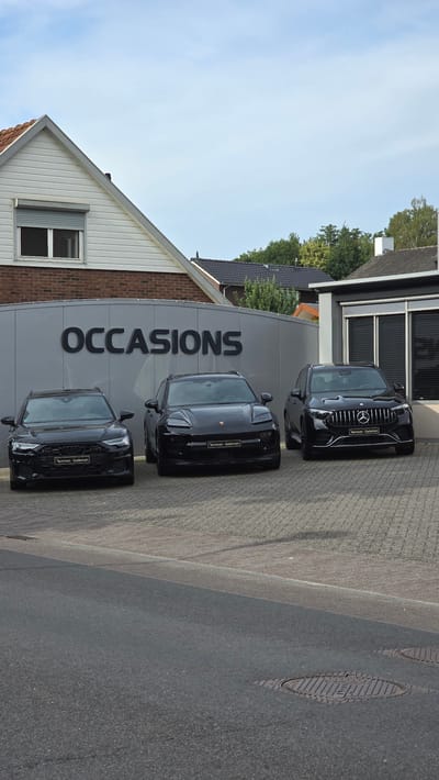 A low-angle, eye-level, outdoor, vertical shot shows three black luxury cars parked in front of a building with the word "OCCASIONS" in large black letters on a grey facade. The cars, from left to right, are an Audi, a Porsche Macan, and a Mercedes-Benz. The Audi and Porsche have license plates that read "Remmen Gademan". The cars are parked on a paved area, with a road in the foreground. The building appears to be a car dealership. The sky is partly cloudy and blue. The overall scene suggests a commercial area, possibly in Zeddam, Netherlands.