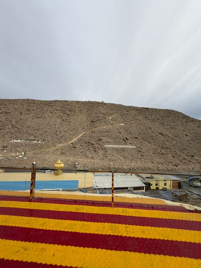 In Chumkar, India, a steep, rocky hillside dominates the scene under a cloudy sky. Large white lettering spells out "CHAKRALA" in the lower left quadrant of the hillside. Further up and to the right, another set of white lettering reads "DHAN GURUNANAKDEVJI". A winding, pale yellow path snakes its way up the hillside. In the foreground, a vibrant, striped walkway with alternating red and yellow bands runs horizontally. Below this walkway, a row of buildings with various roof colors, including blue and yellow, are visible. A golden domed structure, possibly part of a religious building, stands out among the lower structures. A white vehicle is parked near the golden dome. The overall environment appears to be a mountainous, possibly arid region.
