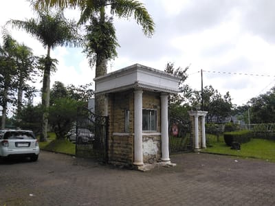 The image displays an entrance to a property or community in Nathene, Laos, under an overcast sky. A prominent stone-clad guardhouse, featuring a white roofline, white trim, and two front-facing white columns, stands centrally on the right side of a dark gray interlocking brick driveway. A closed, ornate black wrought-iron gate is positioned directly adjacent to the guardhouse, spanning the driveway. A circular red sign with a white symbol, indicating a restriction or prohibition, is affixed to the gate. To the left of the driveway, a white SUV, possibly a Suzuki, is parked, facing inward towards the gate. The area is characterized by lush tropical vegetation, including several tall palm trees with visible epiphytes, various green bushes, and neatly maintained grass verges on both sides of the driveway. In the background, partially obscured by trees, are outlines of residential structures. Overhead, power lines are visible against the cloudy sky. No individuals, actions, or interactions are observable within the scene.