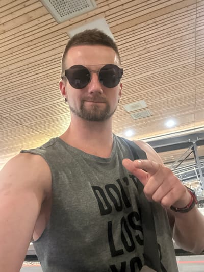 A man is captured in a selfie taken indoors. He is wearing sunglasses and a grey sleeveless t-shirt with black text on it. His left arm is extended towards the camera, with his index finger pointing. He has a short beard and short hair. The background features a wood-paneled ceiling with lighting fixtures and air vents. The overall scene suggests an indoor public space, possibly an airport or transit hub, given the architecture.