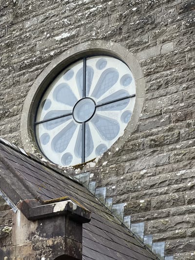 This is a top-down, close-up shot of the exterior of a stone building, likely a church or historical structure, given the architectural details. The primary subject is a large, circular stained-glass window, often referred to as a rose window, which is stylized with a floral or petal-like design. The window is framed by rough-hewn grey stone blocks that form the wall.
Adjacent to the window, and dominating the right side of the frame, is a dark-tiled roof, sloping downwards and away from the viewer. The roof tiles appear to be slate and are weathered, with patches of moss and lichen visible, suggesting an older structure. The stone work of the wall is also weathered, with white and green patches of lichen scattered across its surface, indicating exposure to the elements over time.
The stonework is laid in horizontal courses, with individual blocks having a rough texture and visible mortar lines. The window itself is a prominent feature, with a central circular pane surrounded by radiating, petal-shaped panes of glass. These panes are a light, translucent grey with a subtle pattern etched into them, possibly representing leaded glasswork or a frosted effect. The glass has a reflective quality, showing some of the surrounding architectural elements.
The scene is lit by natural light, casting subtle shadows that highlight the textures of the stone and the roof tiles. The time of day appears to be daytime, likely morning or afternoon, given the brightness and the angle of the light. There are no people visible in the frame, and the focus is entirely on the architectural elements. The overall impression is one of age, solidity, and a sense of place, consistent with historical buildings found in locations like Mountcharles, Ireland. No visible text is present in the image.