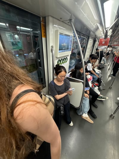 An indoor, eye-level medium shot shows the interior of a subway car during the daytime, with passengers seated and standing. The scene is brightly lit by overhead lights and light coming from outside windows.
In the foreground, the arm and long, light brown hair of a person are visible on the left side, partially obscuring the view of other passengers.
The main focus is on a group of passengers seated on a bench in the middle of the car. On the left, a woman with glasses and dark hair pulled back is looking at her phone. She is wearing a dark grey t-shirt and has a camera with a lens attached hanging from her neck, with a beige and brown plaid bag resting on her lap. Beside her, a person with dark hair is also looking at their phone. Further down the bench, a man with black hair and a white t-shirt is seen from the side, also engrossed in his phone, wearing blue jeans and dark shoes. Next to him, a person wearing a black vest over a white t-shirt and black pants is looking down.
Other passengers are visible in the background, including a person in a black outfit standing and holding onto a metal pole, and another person in a red shirt standing further down the aisle. The subway car interior features white walls, metal poles for support, and windows with reflections showing the outside environment. There are screens mounted on the wall displaying information and advertisements. Signs with Chinese characters are also present, one yellow and triangular with Chinese writing and another red square sign with a QR code. The floor of the subway car is a dark grey material. The overall atmosphere appears to be one of quiet commuting, with most passengers focused on their electronic devices. The image was captured in Xuhui District, China.