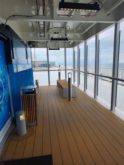 The image shows an indoor walkway with large windows overlooking the sea. The walkway has a wooden floor and a long bench with a silver-colored bollard in front of it. To the left, there is a large blue advertisement panel, a trash can, and a cylindrical light fixture. The ceiling is white and has exposed conduits and lighting fixtures. Through the windows, the sea is visible, as well as the deck of a ship and some people. The overall impression is a modern and clean space with a view of the water.