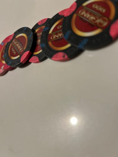 Five hundred-dollar casino chips from The Row complex are displayed on a reflective, light-colored surface in Reno, Nevada, United States. Each chip is primarily black with a central red field, a gold inner ring, and the inscriptions "THE ROW", "GRAND", and "$100". The chips also feature distinctive pink/magenta and blue markings along their edges. They are arranged in a diagonal, partially overlapping line, extending from the lower-left towards the upper-right of the frame. A bright, localized reflection of an artificial light source is present on the surface in the lower-right quadrant. No individuals or specific actions are depicted.