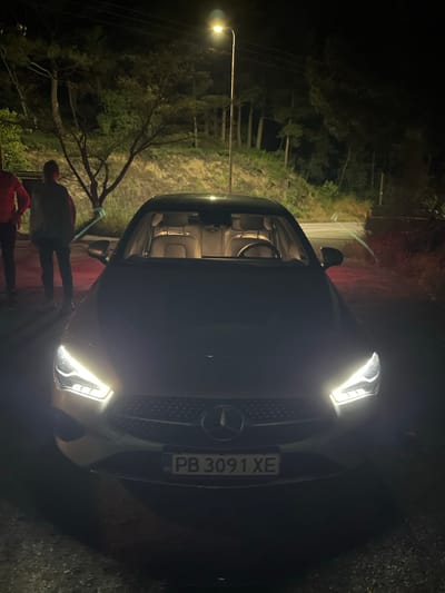 At night, a Mercedes-Benz car with the license plate "PB 3091 XE" is parked on a road, illuminated by its headlights. The car appears to be near or within a wooded area, indicated by trees and foliage. A streetlight further illuminates the area. Two people are standing near the front of the car. The location is presumed to be in Xanthi, Greece, based on information provided. The overall scene presents a nighttime environment on a road near wooded terrain, with a focus on the vehicle and surrounding infrastructure.