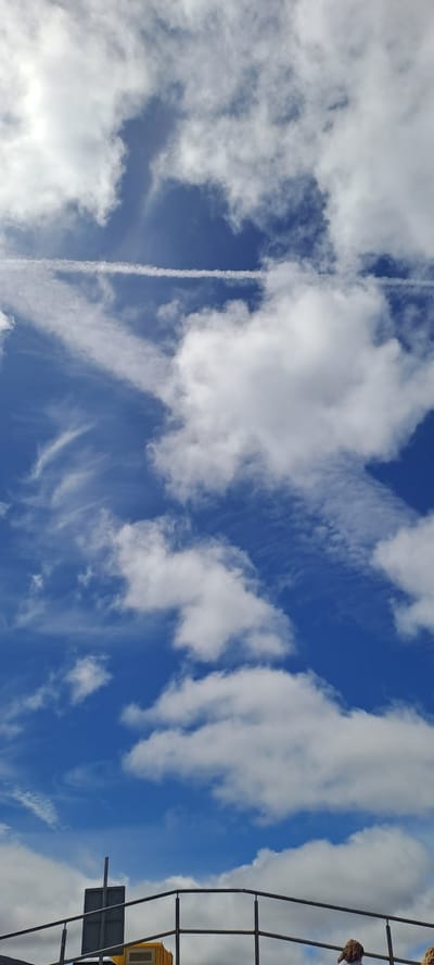 The image shows a bright blue sky with scattered white clouds. A prominent contrail stretches vertically down the center of the frame, suggesting an airplane has recently passed. The clouds are a mix of fluffy cumulus formations and wispy cirrus clouds, indicating fair weather conditions with some atmospheric activity.
On the right side of the image, a dark metal structure, possibly part of a railing or a larger piece of equipment, is visible. A yellow object, possibly a warning sign or a part of a structure, is also present within this framework. A person, wearing a blue and white striped garment, is partially visible in the upper right corner, looking upwards towards the sky.
The image appears to be taken during the daytime, given the brightness of the sky and the clarity of the clouds. The perspective suggests the photo was taken from a slightly elevated position, looking upwards. The presence of the metal structure and the person may indicate a public space or a viewing platform. No visible text is present in the image. The overall mood is serene and expansive, focused on the natural beauty of the sky.