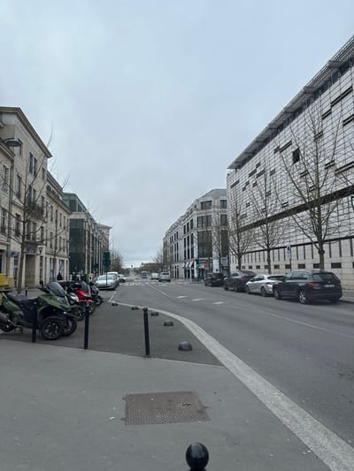 The image presents an urban street view in Serris, France, observed from a low angle looking eastward down a wide, multi-lane road under an overcast sky.

The upper approximately 40% of the frame is occupied by a uniformly light grey, cloud-covered sky.

On the left side, a grey concrete sidewalk occupies the foreground, featuring a square metal manhole cover and a partially visible black spherical bollard at the bottom center. Adjacent to the sidewalk is a paved parking area containing four parked motor scooters: one dark green three-wheeled scooter, one red scooter, and two additional dark-colored scooters. Black cylindrical and spherical bollards are arrayed along the edge of this parking area. Behind these, a multi-story building with a light-toned facade, multiple windows, and small balconies is visible. A blue "La Poste" sign is present on its ground floor. Further down the street, another building displays a partially legible sign, reading "UNIVERSITE & SALLE". Bare deciduous trees, characteristic of winter or early spring, line the sidewalk in front of these structures. One person is partially visible walking on the sidewalk near the "La Poste" building.

The central portion of the image is occupied by an asphalt roadway, featuring a white solid line separating a parking lane from a primary traffic lane on the right. White forward-pointing arrows are painted on the road surface. In the distance, several vehicles, including a white van, are observed traveling away from the viewer.

On the right side of the street, multiple vehicles are parked parallel to the curb, including a black Skoda Kodiaq SUV (with a partially visible French license plate), a silver sedan, and a dark grey sedan/SUV. The building immediately adjacent to the right side of the road is a multi-story, modern structure with a light-colored, grid-patterned facade and numerous windows. Structures consistent with solar panels are visible along its roofline. Bare deciduous trees are situated between this building and the parked vehicles. Further down the street, another multi-story building with a lighter facade and dark-framed windows features a green illuminated sign with the text "SQUARQ" on its ground level.

The scene is devoid of prominent human activity or interactions, focusing on the urban infrastructure and parked/moving vehicles.
