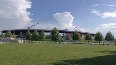 This is a wide shot of the NV Arena in Sankt Pölten, Austria, a football stadium. The stadium, with its distinctive circular roof and external support structures, is seen from across a large, well-maintained green lawn. Several trees are planted in a row between the lawn and the stadium. The sky is blue with scattered white clouds, and the lighting suggests it is daytime, likely late afternoon, given the shadows. In the foreground, the grassy area appears to be a park, with some low concrete structures decorated with graffiti on the left side. There are several people visible on the lawn. On the far left, a few individuals are interacting with the graffiti-covered structures. Towards the right side of the frame, a few people are walking and standing on the lawn, and one person appears to be running. The overall scene depicts a public space adjacent to a sports venue on a clear day.