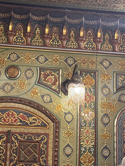 A patterned ceiling features intricate designs in gold, green, and dark blue, reminiscent of Islamic art. Symmetrical floral motifs and geometric shapes are repeated throughout the surface. A decorative lamp fixture, emanating a bright light, is mounted to the ceiling, positioned near the center of the view. The lighting illuminates the detailed textures and ornamental features.