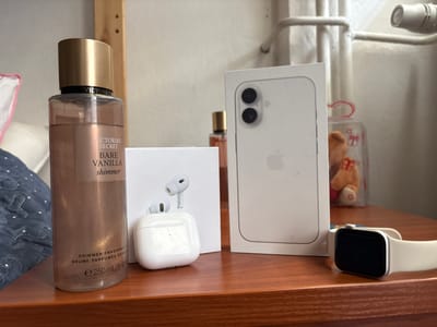 The image displays a collection of personal items arranged on a wooden surface, possibly a dresser or shelf. The primary objects are a tall bottle of Victoria's Secret Bare Vanilla Shimmer body mist, a white charging case for wireless earbuds with two earbuds placed on top, and a white box for an Apple iPhone. To the right of the iPhone box, a white Apple Watch with a light-colored band is visible. In the background, out of focus, a small teddy bear in a clear container with red ribbons and stars can be seen, along with some white shelving or railing. The lighting suggests an indoor setting, possibly during the daytime, with soft light illuminating the objects. The overall arrangement appears to be a display of personal accessories and electronics. Visible text includes "VICTORIA'S SECRET", "BARE VANILLA shimmer", and "SHIMMER FRAGRANCE BRUME PARFUMÉE SCINTILL e 250mL/8.4 flo" on the body mist bottle.