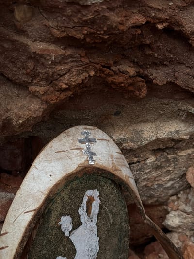 An extreme close-up shot captures a portion of an old, circular object nestled amidst rough, textured rock formations. The object, possibly a lid or part of a vessel, has a worn, light-colored rim, appearing off-white or pale brown, with some vertical scratches or marks. On this rim, a distinct cross-like symbol is visible, formed from several small, reflective, rectangular pieces, possibly metal or tape, suggesting either a repair or a decorative/symbolic addition.
The interior of the circular object is a dark, speckled surface, from which patches of white material are flaking off, revealing the darker layer beneath. Extending from the upper right corner of the frame, a thin, dark, curved piece of material, possibly wood or metal, appears to be part of or associated with the main object, nearly touching its rim.
The background is dominated by highly textured, earthy or rocky surfaces, primarily in shades of dark brown, reddish-brown, and some lighter greyish-brown tones. These surfaces exhibit deep cracks, rough edges, and irregular contours, indicating a natural or rustic setting. No discernible activity or event is taking place, presenting a static, detailed view of the object and its immediate surroundings. The lighting suggests daytime, but without direct sunlight, leaving the time of day and weather indeterminate. No legible text is visible in the image. Given the location context of Staroe Rakomo, Russia, the object could be a historical or archaeological artifact found in a natural or ancient site.