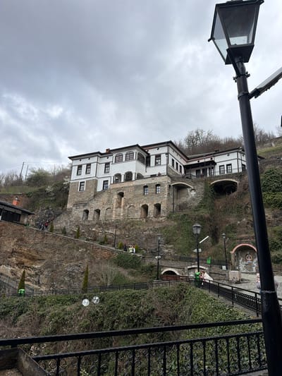 The image captures a multi-story building built into a hillside, potentially a historic or cultural site. The building is white with dark-framed windows, and its façade is partially constructed with exposed stone. The architecture is characteristic of a traditional style, possibly Balkan. The structure is situated on a slope covered in vegetation, including ivy and various shrubs. A path or walkway is visible, edged with a black metal railing. Street lights line the walkway. The sky is overcast. Given the building's style and location, it's very possible this is a building in Kriva Palanka, North Macedonia.
