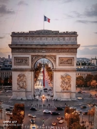 The image shows the Arc de Triomphe in Paris, France, at dusk. The monument is centrally located, with a French flag flying at its top. A street lined with trees and traffic, including cars and a bus, is visible through the archway. People are gathered around the monument and on the surrounding streets, and vehicles are moving in a circular pattern around the base of the Arc. The sky is overcast, with shades of grey and purple. The text "OPPO A17 2026/07/26 22:13" is superimposed in the lower left corner of the image, indicating it is a digital photograph taken with an Oppo A17 phone on July 26, 2026, at 10:13 PM. The city of Құмөзек, Kazakhstan is not depicted in this image.