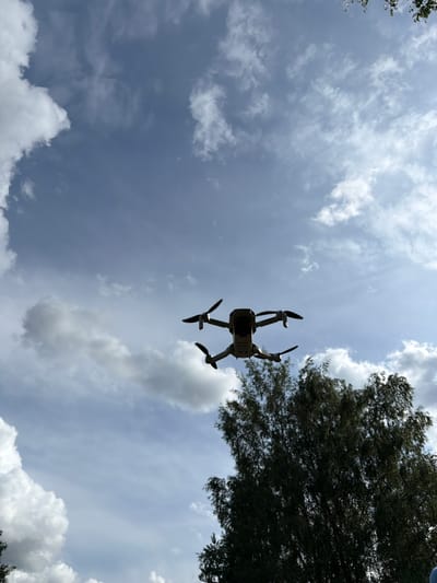 An aerial view captures a drone in flight against a backdrop of a partly cloudy sky and the upper branches of a dark green tree. The drone, silhouetted against the sky, has four propellers and is positioned in the upper center of the frame. The sky is a mix of light blue and scattered white clouds, with a slightly darker hue suggesting it is daytime. The tree foliage, dense and dark, occupies the lower right portion of the image. There is no visible indication of the specific location within Tēraudi, Latvia.