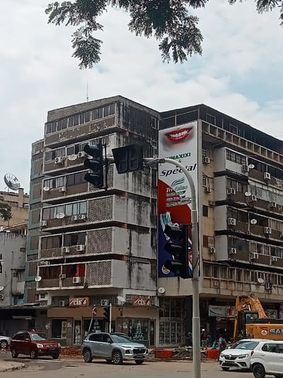 An urban scene in Luanda, Angola, features a multi-story concrete building under an overcast sky, with lush green tree branches overhanging at the top. The building, likely residential or mixed-use, shows signs of wear with stained and weathered facades. It has numerous windows, balconies with patterned railings, and external air conditioning units attached to nearly every floor, along with several satellite dishes.
A large billboard is prominently displayed on the right side of the building. The top portion features a bright, smiling mouth with white teeth, advertising what appears to be a dental product. Below this, the text "KINAXIXI" and "special" are visible, followed by "Tri-Ativo", "Dentes Fortes", "Hálito Fresco", and "Proteção Branca". A large image of a toothbrush is also part of the advertisement.
In the foreground, a busy street is visible with vehicles. A red SUV and a silver SUV are parked or driving on the left side of the road, while a white car is on the right. A set of traffic lights on a pole stands in the center-right, partially obscuring the billboard. At the street level of the main building, two shopfronts are visible, both displaying the text "CIN" with a logo.
To the right, there is evidence of ongoing construction or roadwork, with an orange excavator clearly visible, marked with "SANY". A few people can be seen near the construction site. The overall setting is a typical daytime urban environment, bustling with activity, architectural details, and commercial advertisements. Another sign near the construction reads "ANIVERSÁRIO". Vehicle license plates, such as "LD-12-99-FH" and "LD-95-58-FL", indicate the local context.
