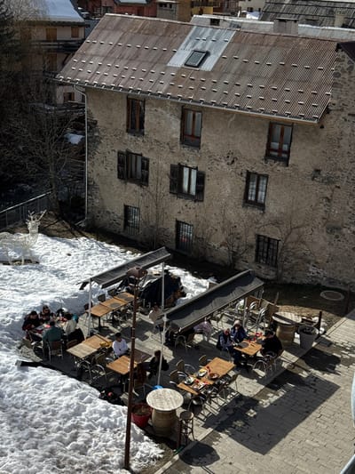 This image depicts an outdoor patio area adjacent to a multi-story stone building in La Salle-les-Alpes, France. The building features a rough, light-brown stone facade with several windows, some on the lower levels equipped with closed dark wooden shutters. The upper windows lack shutters. The roof is corrugated metal, predominantly a weathered brown, with a distinct, newer-looking silver corrugated section and a single skylight. A white rain gutter and downspout are visible on the left side of the building.
The outdoor patio is partially covered in snow on the left and a paved with light-colored bricks on the right. Approximately 18 to 20 individuals are seated at various rectangular and round wooden tables. Some tables are positioned under dark, retractable awnings supported by metal frames. People are observed eating, drinking, and socializing.
Specific objects include a central brown pole with a light fixture. On the snow-covered ground to the far left, a large, white, illuminated decorative deer figure is present. On the paved section, two wooden barrels are visible, one serving as a table base and another as a decorative element next to a gray planter. A red pot or planter is near the base of the central pole. Bare trees with no leaves are visible in the background on the left and close to the building's facade. The scene is under natural daylight, casting clear shadows from the awnings, tables, and structures onto the paved ground. Further buildings are visible in the distant background.