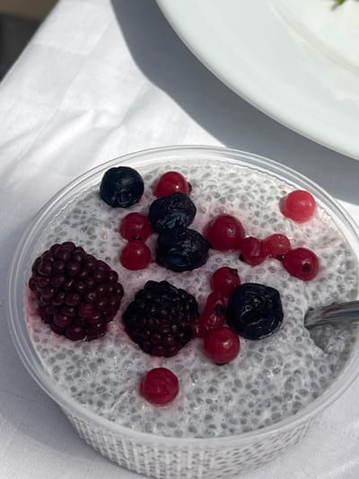 A high-angle shot depicts a clear plastic container filled with chia seed pudding, topped with an assortment of berries. The berries include red currants, blueberries, and blackberries. A spoon rests in the pudding. A white plate is partially visible to the left of the container. The container sits on a white tablecloth. The location is indicated as Szigetszentmiklós, Hungary, though the image contains no information about the location. The scene is a still life, with no people or actions evident.