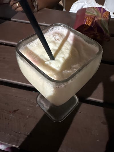 A high-angle photograph depicts a clear, stemmed glass filled with a frothy, opaque, off-white beverage exhibiting subtle pinkish coloration. A black plastic straw is inserted into the drink, angled towards the upper left. Condensation is visible on the exterior of the glass. The glass is positioned on a dark brown, horizontally-slatted wooden table surface. In the upper left background, a human hand is visible, wearing a gold ring on the fourth finger and a metallic wristwatch, resting on the table. In the upper right background, a reddish-purple object patterned with repeating gold pineapple motifs is partially visible, adjacent to a crumpled white fabric. The scene is dimly lit, suggesting an evening or indoor setting. This environment is consistent with a casual dining establishment or bar, a type of venue found in the city of Roatán, Honduras; however, specific geographical identifiers within Roatán are not present in the image.