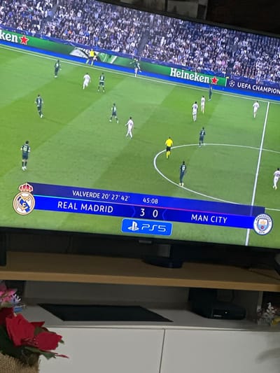 The scene depicts a domestic setting in Lancaster, United Kingdom, focused on a television displaying a football (soccer) match. The television occupies the upper two-thirds of the image, showing a UEFA Champions League fixture between Real Madrid (wearing white kits) and Manchester City (wearing dark green/teal kits). The scoreboard at the bottom of the screen indicates Real Madrid leading Manchester City 3-0, with "VALVERDE 20' 27' 42'" identifying the goal scorer and times. The match time is displayed as "45:08." A PlayStation 5 (PS5) logo is visible on the scoreboard graphic. Numerous players from both teams are distributed across the bright green pitch, along with two referees in yellow. Advertising hoardings visible behind the players display "Heineken" and "UEFA CHAMPIONS." A large, light-clad crowd is visible in the stadium stands.
Below the television, a wooden-toned TV stand with two shelves is present. On the right side of the bottom shelf rests a black electronic device, identifiable as a Sky Q Mini box, with a green indicator light. A second dark, rectangular object is on the left side of the same shelf. In the lower-left foreground, a decorative item with red artificial flowers and a woven base is partially visible. Beneath the TV stand is a white cabinet with two doors.