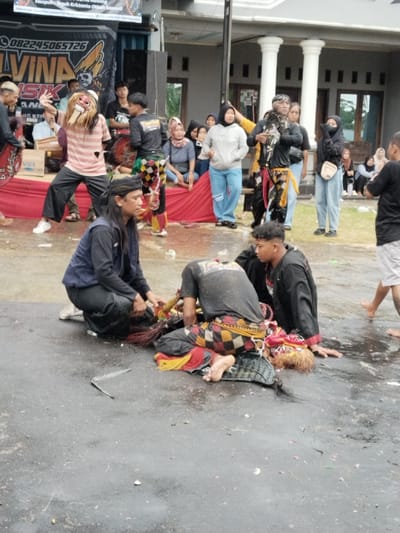 This outdoor scene in Sumberejo, Indonesia, depicts a cultural performance or ritual on a wet, dark gray paved surface. In the foreground, a man with long dark hair, wearing a dark blue vest over a black shirt and black pants, crouches on his knees. He is attending to another man lying prone on the wet ground to his right. This prone individual wears a dark t-shirt and patterned pants with red, yellow, and black checkered motifs, typical of a traditional costume, and a red and yellow headdress is visible near his head. A third young man in a black long-sleeved shirt kneels beside the prone individual. The pavement is wet with puddles and scattered white and pink debris, possibly flower petals.

In the mid-ground, to the left, a performer in a striped shirt and dark pants is actively posing while wearing a large, light-colored mask with prominent teeth and shaggy hair. Other individuals in traditional black and yellow patterned costumes are also present.

The background features a light-colored building with white pillars. A large black banner displaying "ALVINA MUSIK" and a phone number (082245065726) is prominently mounted. A black speaker is visible to the left of the banner. A raised platform, covered with red fabric, serves as a temporary stage or seating area, where an "AQUA" branded water bottle is present. Approximately 15-20 spectators, mostly women in casual modern attire, some wearing hijabs, are observing the event from this platform and surrounding areas.