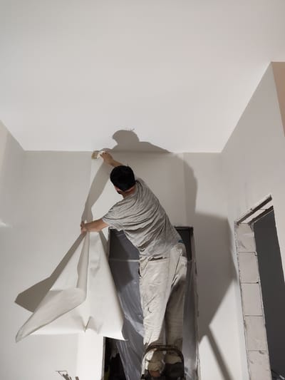 A person is standing on a ladder, applying wallpaper to a wall. The person is wearing a striped t-shirt and light-colored pants, with protective coverings on their shoes. They are holding a roll of wallpaper and using a brush to smooth it onto the wall. The room appears to be under renovation, with unfinished elements visible, such as exposed brickwork around an opening. The walls and ceiling are painted white. The lighting suggests it is daytime, with shadows cast by the person and the objects in the room. The scene is indoors, in what appears to be a residential setting in Skurygino, Russia, during a home improvement project.