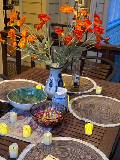The image depicts an outdoor dining table set for a meal. A wooden table is covered with two circular placemats with frayed edges, a small linen runner with text, and a floral arrangement in a vase. In the center of the table is a glass bowl filled with individually wrapped candies, and a green and white striped bowl. A bottle of Corona beer sits on one of the placemats. There are several small, battery-operated candles scattered around the table. In the background, a wooden chair is visible, as is a wall of a building with windows. The lighting suggests it is evening. The location is described as Газета Труд Снт, Russia. No people are present in the image.