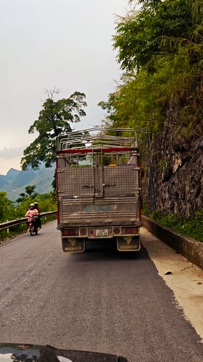 The image depicts a road scene in Cán Tỷ Commune, Vietnam. A medium-sized truck with a cargo bed dominates the center of the frame, driving on a narrow road. Behind the truck, a motorcycle with two riders is approaching. To the right of the truck is a rocky embankment with green vegetation. The road itself appears well-maintained. In the background, a mountainous landscape is visible under a cloudy sky. A distinctive tree stands out against the backdrop.