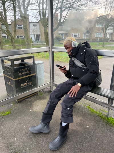 A person sits at a bus stop, legs stretched out, wearing black pants, a black puffer jacket, and black rain boots. They have blonde hair, wear white headphones, and hold a phone. A gray fanny pack is slung across their chest. A gray trash bin labeled "Litter & Dog Waste" sits nearby. Buildings are visible through the bus stop's glass.