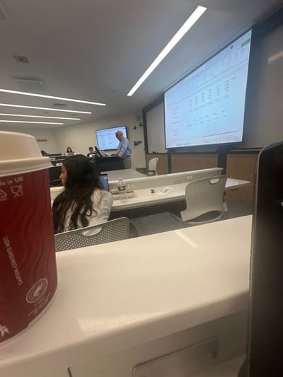 The image captures a modern classroom or lecture hall, likely within a college or university in Wellesley, United States. In the immediate foreground, a red and white paper coffee cup, bearing "CAUTION CONTENTS HOT" and "Made in UK" text, rests on a white desk, partially obscuring the lower left of the frame.

A female student with long dark hair, wearing a white top, is seated in the mid-ground at a light-colored desk with an open laptop, facing the front of the room. A plastic water bottle and a small container with snacks are visible on her desk. Light grey, perforated chairs are used by students.

At the front of the room, an older Caucasian male, wearing a light blue button-down shirt and khaki pants, stands behind a dark grey podium, apparently lecturing or presenting. He is positioned between two projection screens. The large screen on the right displays a Microsoft Excel spreadsheet with multiple columns of numerical data, including month labels such as "Jan," "Feb," "Mar," etc. A smaller screen behind the presenter shows a similar or related spreadsheet.

Several other students are partially visible in the background, seated at identical light-colored desks equipped with visible power outlets. The room is well-lit by multiple linear LED light fixtures recessed into the white ceiling. The walls are predominantly white, with darker panels and wood trim beneath the main projection screen. The floor appears carpeted.