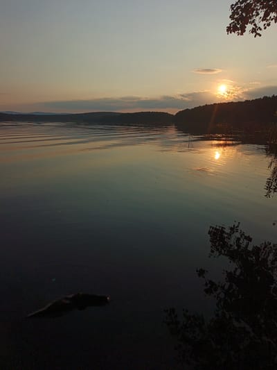 A body of water is depicted at sunset, with the sun partially obscured by clouds and casting a warm glow on the water's surface. The horizon features a silhouetted tree line. In the foreground, a dark object floats on the water. The scene suggests a natural environment, possibly a lake or river, in Nepryakhinskoye Sel'skoye Poseleniye, Russia, during the evening.