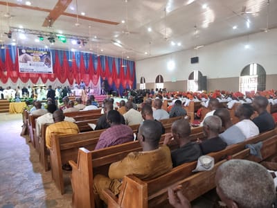 The image shows a wide-angle view of the interior of a church in Ungwan Liman, Nigeria. The church has a high, vaulted ceiling with exposed wooden beams and numerous ceiling fans and lights. The walls are white, and the space is decorated with red and white drapes that hang from the ceiling and frame a stage area at the front. On the stage, there is a large banner advertising an event, a podium, musical instruments, and several people, some of whom appear to be giving a presentation or performing. The main seating area is filled with rows of wooden pews, occupied by a congregation of men and women. Many of the attendees are facing the stage. The floor is tiled. The overall impression is of a religious gathering taking place in a community church.