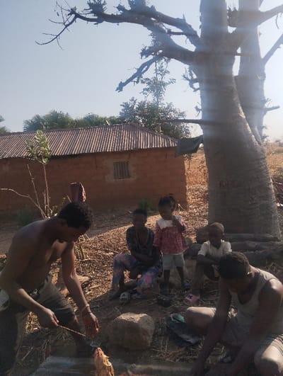 A group of people are gathered outside near a small mud brick house with a corrugated metal roof. A shirtless man is skewering meat over a fire, while another person is sitting next to him. Nearby, a woman sits with her legs crossed, and a young girl in a pink dress and blue shorts stands next to her. A small boy is also sitting near the group. A large tree stands in the background.