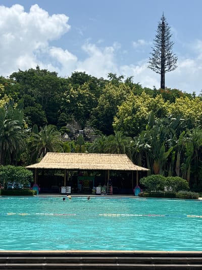 In Yingzhou, China, a swimming pool with turquoise water is situated in front of a dense green forest. A thatched-roof pavilion with wooden posts is positioned behind the pool. Several people are visible in the water. The background features a man-made waterfall and rocks, and beyond that, a tall, slender tower resembling a tree. The sky is blue with scattered white clouds. The foreground shows a dark-colored tiered structure leading down to the pool.