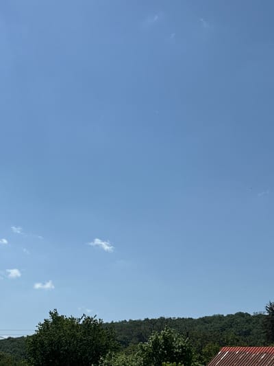 The image presents a landscape with a clear blue sky occupying the majority of the frame. Small, scattered clouds are visible. Along the right edge of the image, there is a line of dense greenery, suggesting a forest or hillside. A section of what appears to be a red, corrugated metal roof is visible at the top right corner. Based on the information provided, the location is Aprilovo, Bulgaria.