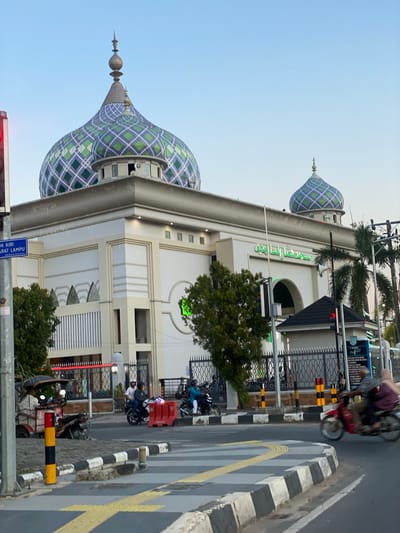The image shows a mosque with a distinctive green and blue diamond-patterned dome, located in a city in Jawa, Indonesia. The mosque is a large, white building with several smaller domes and architectural details. In front of the mosque, there is a street with traffic, including motorcycles and a three-wheeled vehicle. People are visible on the motorcycles, some wearing helmets. The street has crosswalks and traffic barriers. Trees and palm trees are also present, suggesting a warm climate. The sky is clear and blue, indicating daylight. There are also street signs and utility poles visible.