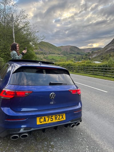 A blue Volkswagen Golf R with license plate CA75 RZX is parked on the side of a road. The vehicle is situated near a metal guardrail separating the road from a grassy hillside. A woman is seen standing near the car, holding a smartphone, possibly taking a photo. The surrounding environment includes rolling hills and mountains under a cloudy sky. The location is Llangynog, United Kingdom.