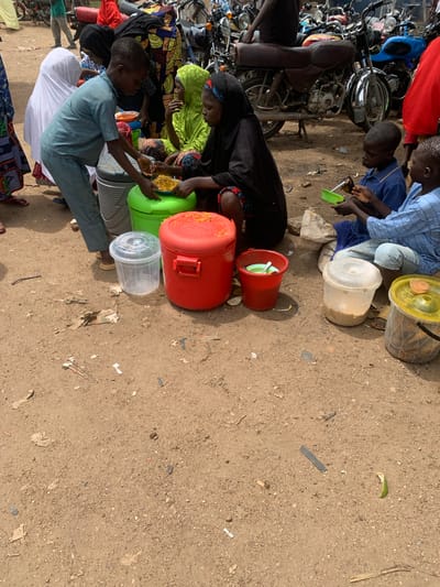 In Project, Nigeria, a group of children and adults are gathered outdoors on a dirt ground, possibly at a market or communal gathering. In the foreground, a young boy in a blue shirt and pants is serving food from a large red container to a child wearing a blue shirt. To the left, a girl in a white hijab stands with a large green container. Several other people in traditional attire are seated on the ground, including a woman in a green headscarf and a woman in a black headscarf. Various containers, including large plastic tubs and buckets, are scattered around the scene. In the background, numerous motorcycles are parked, suggesting a busy area. The environment appears somewhat dusty and unpaved, typical of many areas in Nigeria.