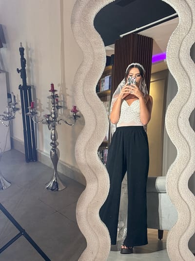 The image is a vertically oriented, full-body mirror selfie taken in an indoor setting, likely a home. The subject, a woman, is wearing a white, sequined top with a sheer overlay and black, wide-leg pants. She has on a sparkling headband and is holding a smartphone in her hands, capturing the reflection. The mirror itself has a textured, fluffy white border with a wavy pattern.
In the reflection, we see the woman from the chest up, holding her phone to take the picture. She appears to be in a room with wooden paneling on the wall behind her, and a hint of purple light from what looks like a shelf or accent lighting. Below her, reflected in the mirror, is a sleek, silver candelabra with several pink candles. There are also reflections of other objects, including what appears to be part of a camera tripod and possibly some books on shelves in the background. The floor in the reflected view is tiled. The lighting suggests it is daytime or well-lit interior. The overall mood is casual and self-aware. The location context provided is Sweileh, Jordan.