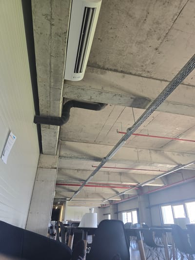 The image shows an interior space with industrial features, possibly a warehouse or storage area, located in Gara Elin Pelin, Bulgaria. The main components are concrete walls and ceilings, with exposed ductwork and piping. An air conditioning unit is mounted on a ledge along the wall. A thick black pipe is also connected to the wall near the AC unit. The floor is a light-colored, possibly linoleum or similar material, and a piece of paper lies on the floor. Overhead, a metal cable tray runs along the ceiling. In the background, stacked office chairs are visible, indicating an office or storage area for office supplies. There are no people visible in the frame.