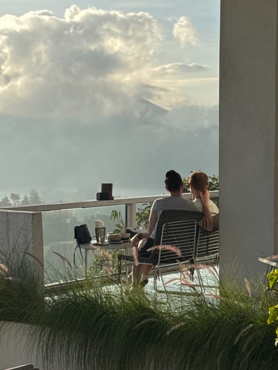 Scene: A cafe in Kedisan, Indonesia, viewed through a glass-enclosed balcony.

People: Two individuals are present. One is a young boy with reddish hair, leaning towards another person, presumably engaged in conversation. The other person, possibly an adult, is wearing a grey shirt.

Objects: Chairs and tables are arranged on the balcony. A purse, cup, and possibly other items are on the table.

Environment: The scene is outdoors, with a view of cloudy sky. Pampas grass is near the right edge of the balcony.