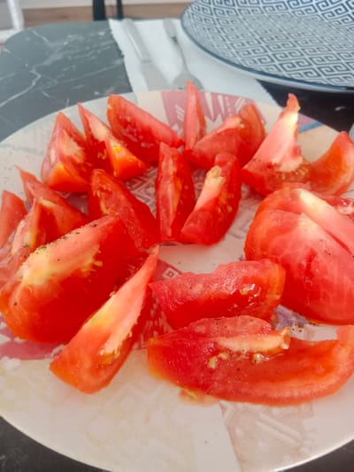 This media file shows a close-up of sliced tomatoes on a plate. The tomatoes are cut into wedges and appear to be seasoned with salt and pepper. They are a vibrant red color with some lighter yellow flesh visible. The plate has a patterned rim with abstract designs in shades of pink, white, and gray.

In the background, a portion of a table with a dark, marbled surface is visible. A white napkin and part of a fork or knife can be seen behind the plate, suggesting a meal setting. A blue and white patterned plate is also visible in the upper right corner of the image, adding to the impression of a dining environment. The lighting suggests it is daytime, possibly indoors, as there are no strong shadows indicative of direct sunlight. The overall scene conveys a sense of simple, fresh food preparation. There is no visible text in the image.