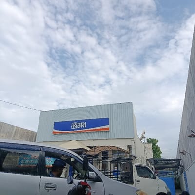 The image shows a view of a bank building with a "BRI" sign in Kecamatan Asembagus, Indonesia, located at coordinates -7.75 latitude and 114.225 longitude. The building is constructed of white corrugated metal panels, and the sign has a blue and orange background. A white van is parked in front of the building. A man in a blue shirt is sitting in a silver car parked in the foreground. There are multiple white canopies above the building. The background is a partly cloudy sky with white clouds against a light blue backdrop. The scene appears to be on a street in a urban or suburban area.