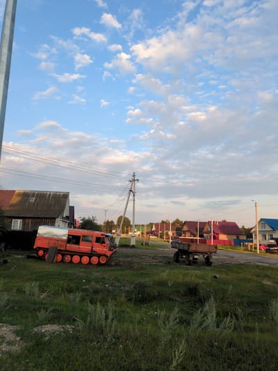 The image captures a scene in the city of Molochnaya Ferma, Russia, under a bright, cloudy sky. In the foreground, a red tracked vehicle, possibly a snowcat or an all-terrain vehicle, is parked on the grass. The vehicle has multiple wheels visible along its sides. To its right, a wooden trailer with four wheels is also present.
The background reveals a street lined with various houses, characterized by their diverse roof colors, including red, brown, and blue. Power lines and poles are visible throughout the scene, indicating the presence of infrastructure. Trees are scattered among the houses and along the street. The overall environment appears to be a rural or semi-rural residential area. No people are visible in the image.