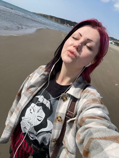 The image displays a young adult female captured in a high-angle selfie on a sandy beach in Ulcinj, Montenegro. She has closed eyes and puckered lips, facing slightly right of center. Her hair is styled with a prominent split dye, dark on the left side and magenta on the right. White wired earphones are inserted into both ears, with the cord draped across her upper chest. She is wearing a grey, white, and brown plaid shirt-jacket, unbuttoned, over a black t-shirt featuring a distressed white and grey graphic print of a stylized female face holding an apple. A brown leather strap from a cross-body bag is visible over her right shoulder. Partially visible below her t-shirt are red and black plaid trousers or skirt. The immediate foreground consists of dark, wet sand with gentle ocean waves receding. In the background, the sea extends to a distant coastline marked by light-colored cliffs and hills under a clear blue sky. Faint white structures are visible on the far right along the distant shoreline.
