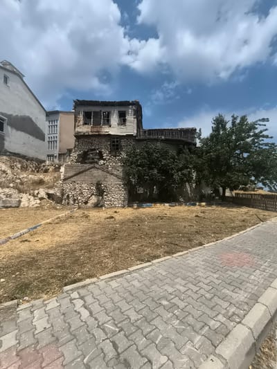 The image depicts an abandoned, multi-story building with a stone foundation and weathered wooden upper floors, situated in a dry, grassy area. The building appears structurally unsound, with damaged roofing and boarded-up windows. Adjacent to it, a modern, multi-story building with a light-colored facade and visible windows stands on a higher elevation. The foreground shows a paved walkway with interlocking gray pavers, bordered by a curb. The background includes a large, mature tree, a wooden fence, and a blue sky with scattered clouds. A portion of the area around the abandoned building is covered in dry, straw-like vegetation. The scene appears to be in a semi-urban or historical district. The location is identified as Eğirdir, Turkey.
