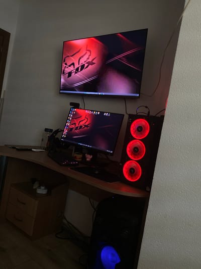 The image shows a computer setup in a room. There is a large flat screen television mounted on the wall to the left. Next to it is a computer monitor with a black bezel, showing a desktop background. A keyboard is visible on the desk along with what appears to be a computer tower. The computer tower has three illuminated fans with red lights. In the far right of the image, there is another computer with a fan illuminated in blue. The image doesn't show any indication of the location being in Sklave, Bulgaria or any other location. The room appears to be indoors with a carpeted floor. A small wooden cabinet is visible below the computer.